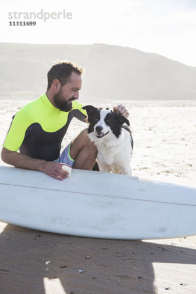 Mann mit Hund  der Paraffin auf sein Surfbrett am Strand legt.