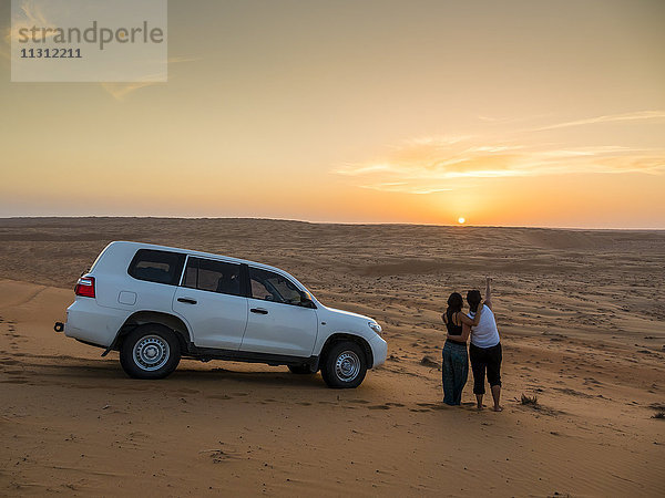 Oman  Al Raka  zwei junge Frauen stehen neben einem Geländewagen auf einer Wüstendüne und beobachten den Sonnenuntergang.