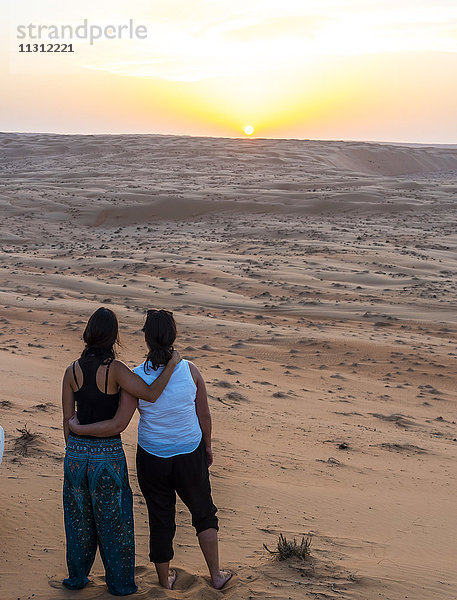 Oman  Al Raka  zwei junge Frauen  die Arm in Arm auf einer Wüstendüne stehen und den Sonnenuntergang beobachten.