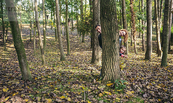 Eltern und kleine Tochter verstecken sich hinter einem Baumstamm