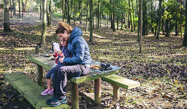 Mutter und Tochter sitzen am Picknickplatz im Wald und spielen mit dem Smartphone.