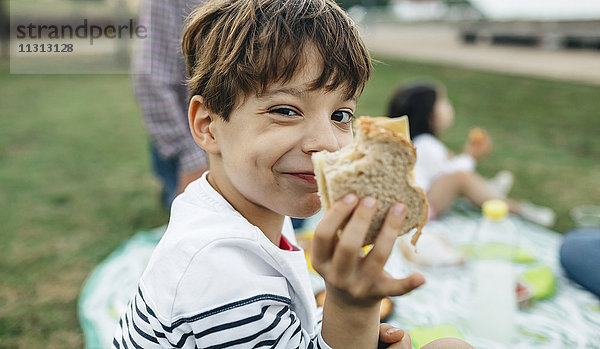 Porträt eines lächelnden Jungen  der ein Sandwich mit seiner Familie im Hintergrund hält.