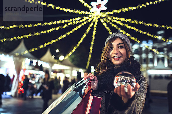 Porträt einer glücklichen Frau mit Taschen und Schneekugel auf dem Weihnachtsmarkt