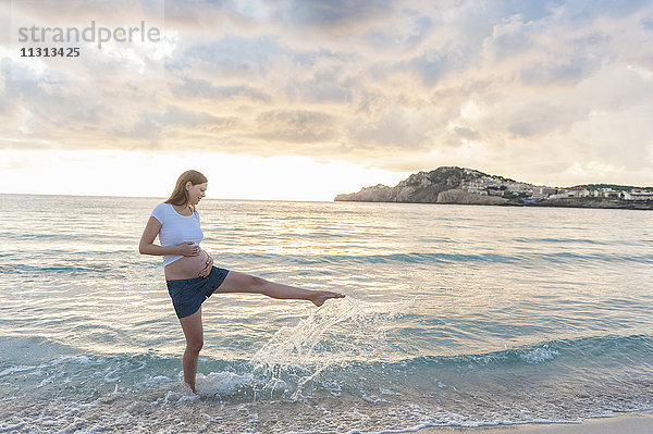Glückliche schwangere Frau am Strand  die mit Wasser spritzt.