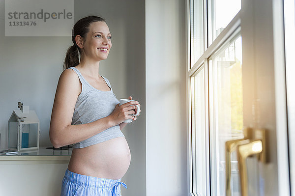 Lächelnde schwangere Frau hält Tasse am Fenster