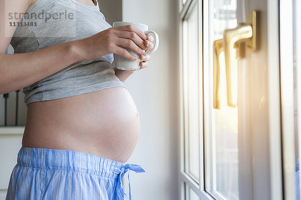 Nahaufnahme einer schwangeren Frau mit einer Tasse am Fenster