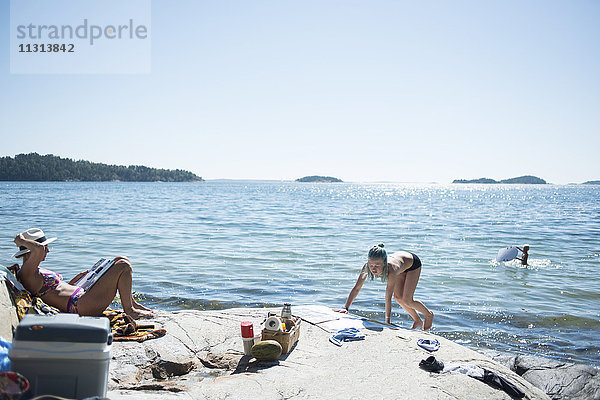 Frauen beim Sonnenbaden am See mit einem spielenden Jungen im Hintergrund