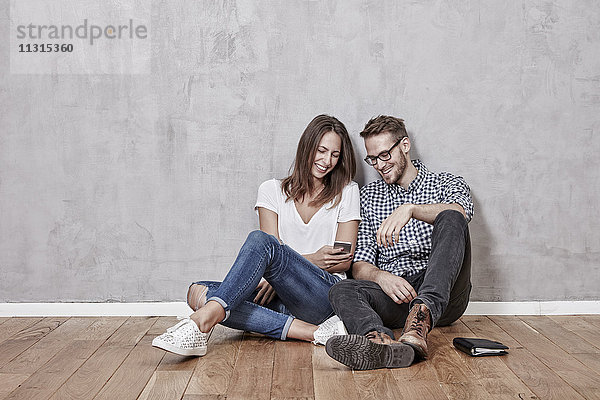 Lächelndes junges Paar auf dem Boden sitzend mit Handy