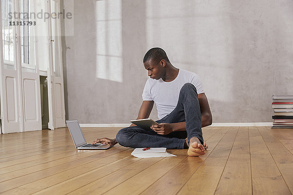 Junger Mann auf dem Boden sitzend mit Laptop und Tablett