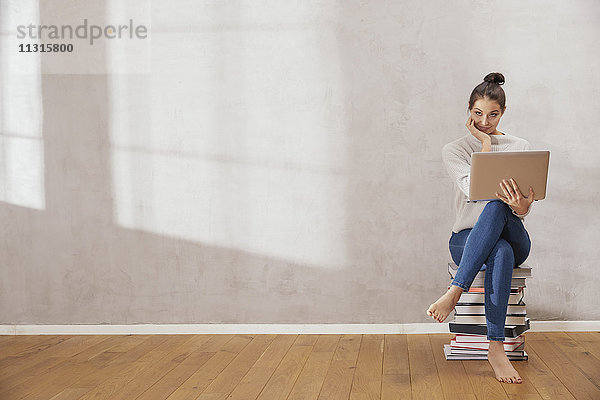 Lächelnde junge Frau sitzt auf einem Bücherstapel mit Laptop