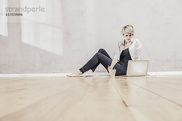 Frau auf dem Boden sitzend mit Kaffeetasse und Laptop