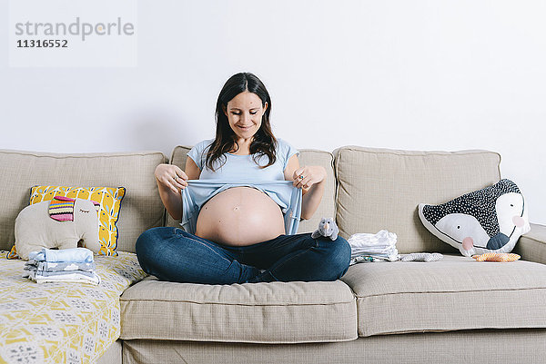 Schwangere Frau sitzt auf der Couch und zeigt den Bauch.