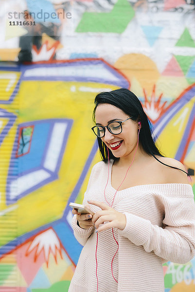 Porträt einer glücklichen jungen Frau mit Kopfhörer und Handy vor der Graffiti-Wand