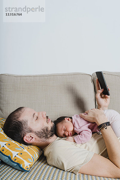 Vater auf der Couch liegend mit neugeborenem Mädchen über Smartphone