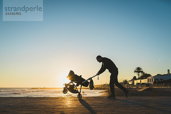 Mann  der bei Sonnenuntergang mit einem Kinderwagen am Strand spazieren geht.