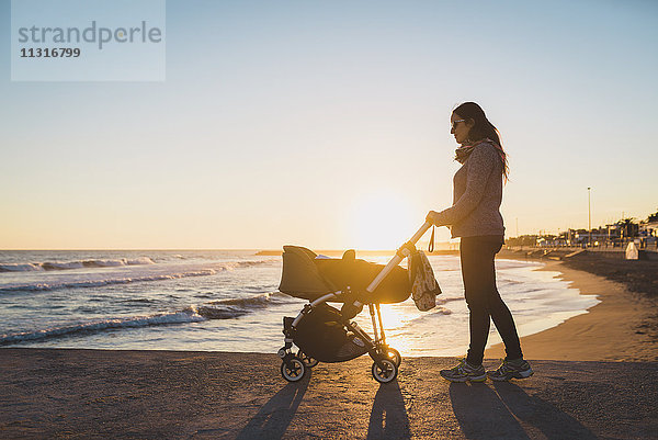 Frau  die bei Sonnenuntergang mit einem Kinderwagen am Strand spazieren geht.