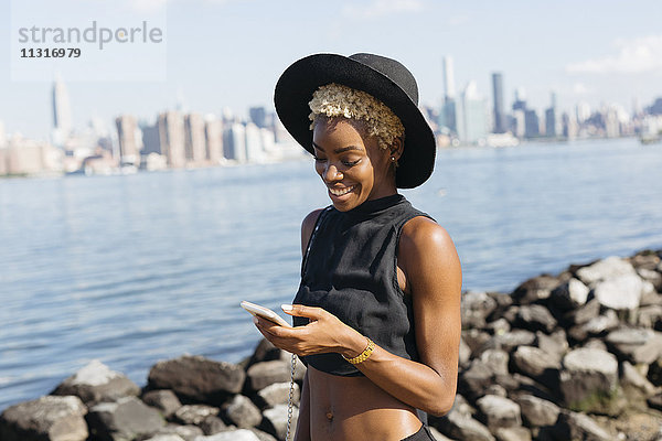 USA  New York City  Brooklyn  lächelnde junge Frau am East River mit Blick aufs Handy