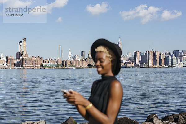 USA  New York City  Brooklyn  lächelnde junge Frau am East River mit Manhattan-Skyline im Hintergrund