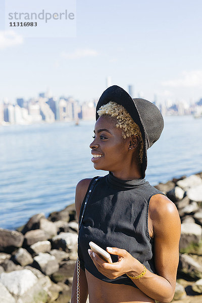 USA  New York City  Brooklyn  lächelnde junge Frau am East River mit Handy