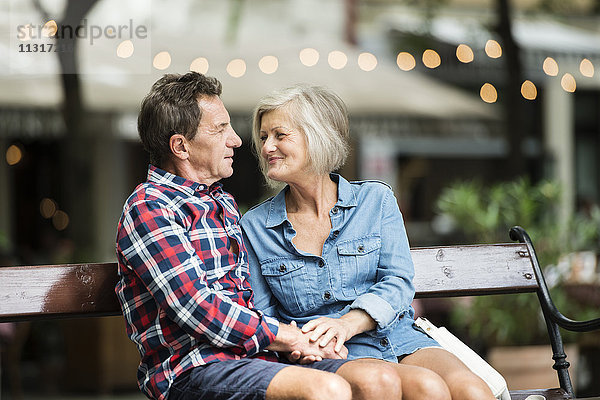 Seniorenpaar sitzt zusammen auf einer Bank und hält sich an den Händen.