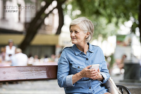 Seniorin mit Smartphone sitzt auf der Bank in der Stadt