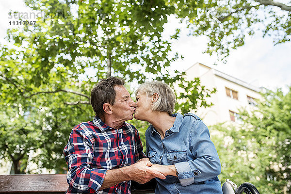Seniorenpaar  das sich auf der Bank küsst und Händchen hält.