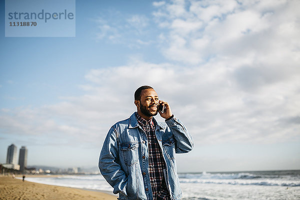 Spanien  Barcelona  lächelnder junger Mann mit Handy am Strand