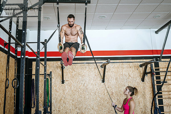 Mann bei Übungen an Ringen im Fitnessstudio mit Frau als Assistentin