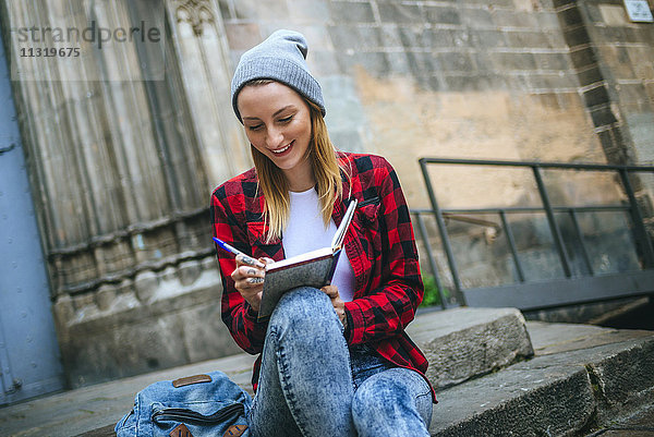 Spanien  Barcelona  lächelnde junge Frau sitzt auf einer Treppe und schreibt in ein Notizbuch.