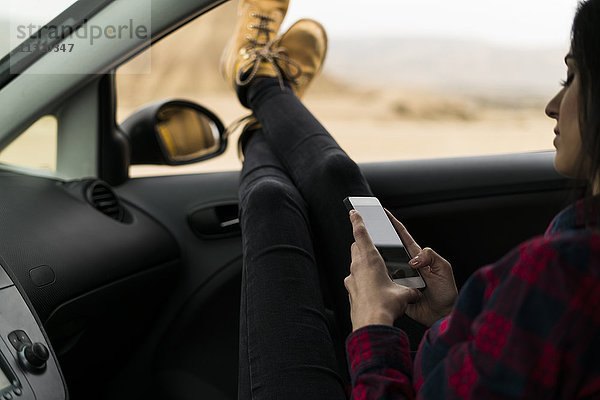 Beine einer jungen Frau  die sich aus dem Autofenster lehnt  während sie ihr Handy benutzt.