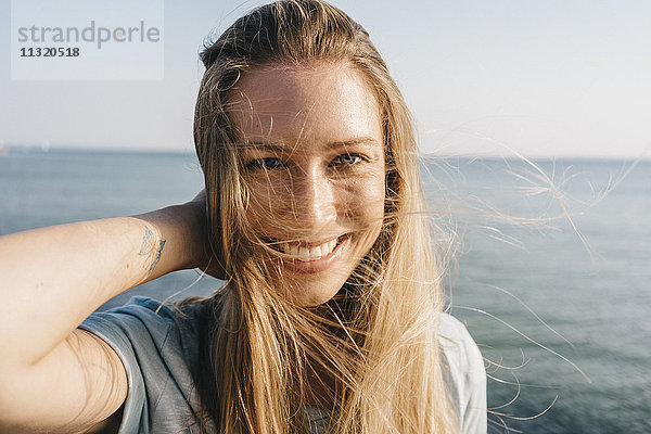Porträt einer glücklichen jungen Frau mit blasenden Haaren vor dem Meer