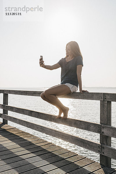 Junge Frau sitzt am Steg bei Gegenlicht und nimmt Selfie mit Handy mit