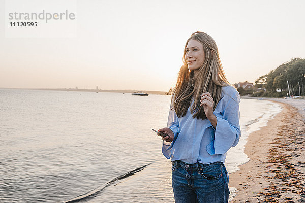 Lächelnde junge Frau mit Smartphone am Strand stehend