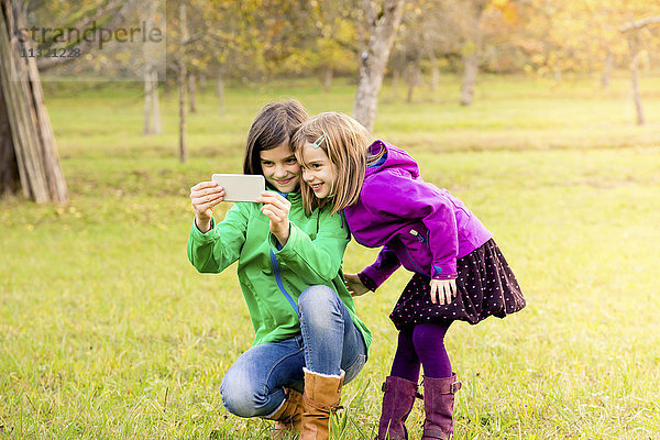 Zwei lächelnde Schwestern nehmen Selfie mit Handy im Herbst auf einer Wiese mit.