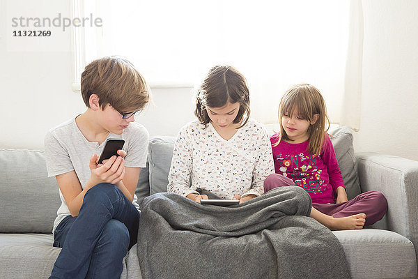 Mädchen sitzend auf der Couch mit Mini-Tablette  während Bruder und Schwester sie beobachten
