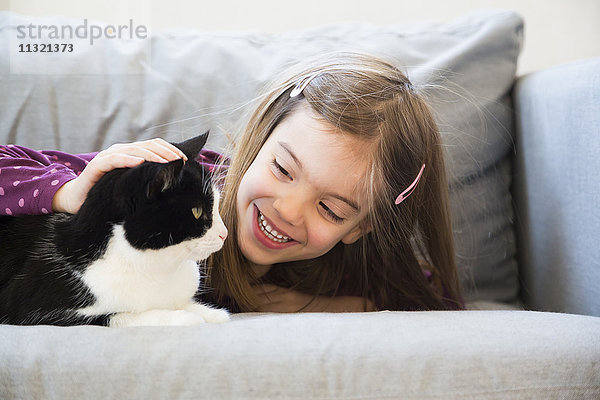 Porträt eines glücklichen kleinen Mädchens  das auf der Couch liegt und die Katze streichelt.