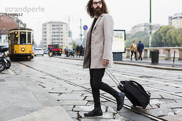 Stilvoller junger Mann mit kofferüberquerender Straßenbahn