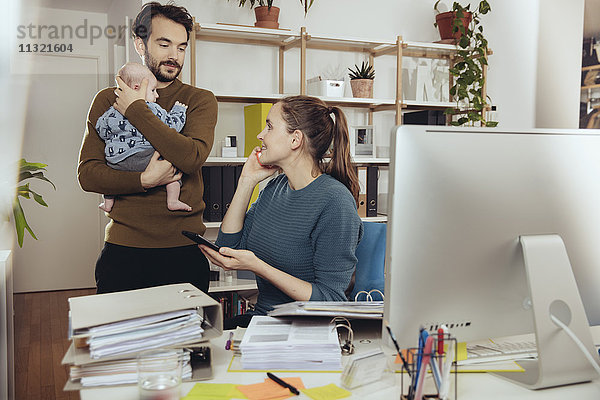 Lächelnde Mutter am Schreibtisch mit Blick auf Vater mit Baby im Home-Office