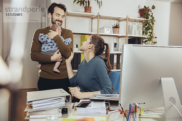 Lächelnde Mutter am Schreibtisch mit Blick auf Vater mit Baby im Home-Office
