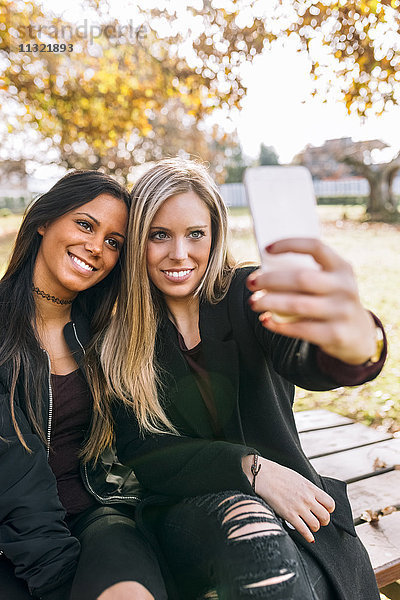 Zwei lächelnde junge Frauen auf der Parkbank mit einem Selfie