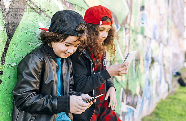 Zwei Kinder lehnen sich mit Hilfe von Smartphones an die Graffiti-Wand.