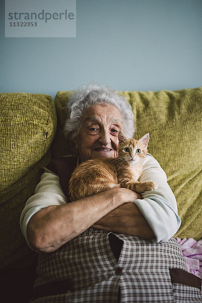 Porträt einer glücklichen Seniorin  die mit ihrer Katze auf der Couch kuschelt.