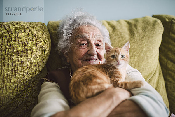 Porträt einer glücklichen Seniorin  die mit ihrer Katze auf der Couch kuschelt.