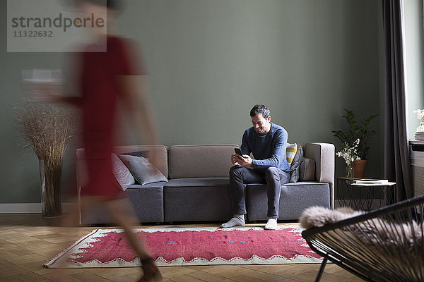Mann sitzt auf der Couch im Wohnzimmer  während seine Freundin im Vordergrund vorbeigeht.