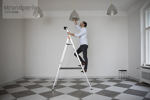 Mann steht auf einer Leiter in einem leeren Raum und wechselt die Glühbirne der Deckenleuchte.