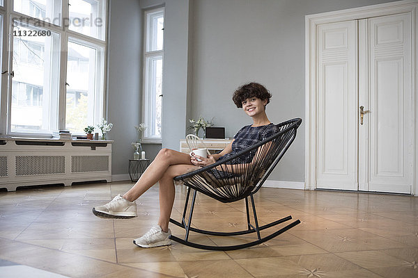 Porträt einer lächelnden jungen Frau  die zu Hause auf einem Schaukelstuhl sitzt.