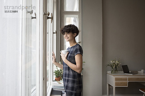 Porträt einer lächelnden jungen Frau  die zu Hause durchs Fenster schaut.