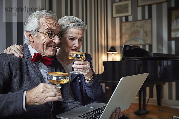 Fröhliches Seniorenpaar trinkt Champagner und benutzt den Laptop zu Hause.
