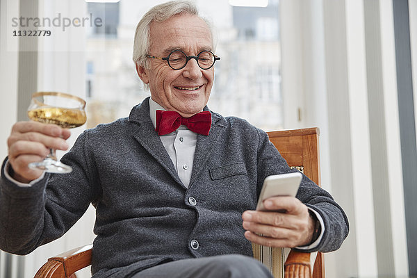 Lächelnder älterer Mann mit Handy auf Stuhl sitzend mit Champagnerglas
