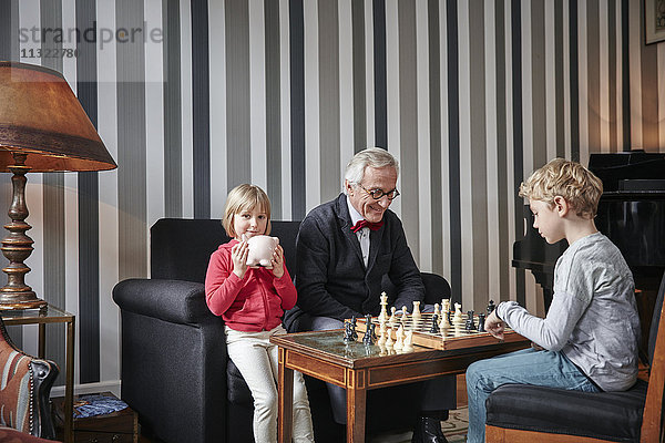Großvater und Enkel beim Schachspielen im Wohnzimmer mit einem Mädchen neben ihnen